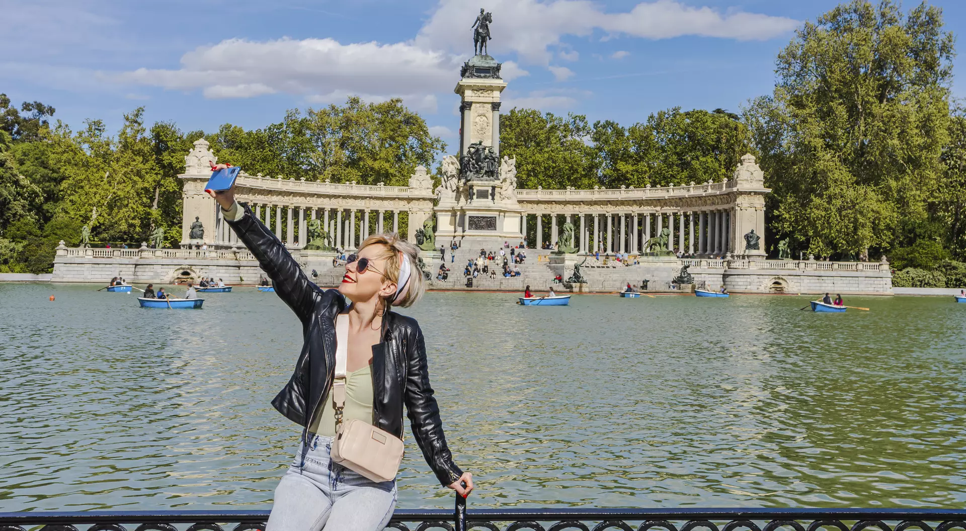 A person taking a selfie by a lake with a monument and trees in the background on a sunny day.
