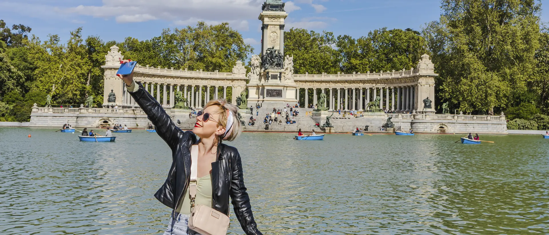 A person taking a selfie by a lake with a monument and trees in the background on a sunny day.