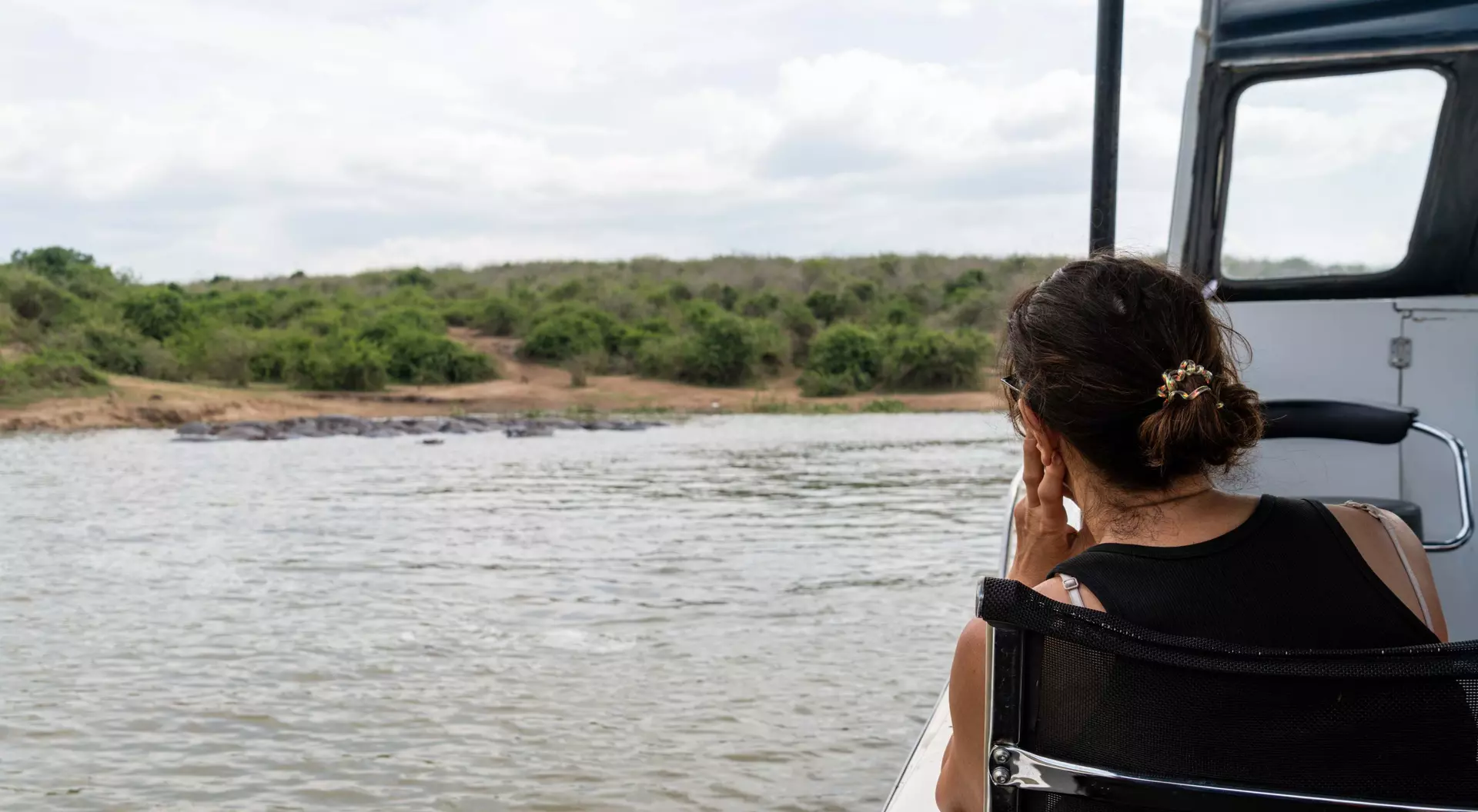 A person sitting on a boat, gazing thoughtfully at the water and surrounding greenery under a cloudy sky.