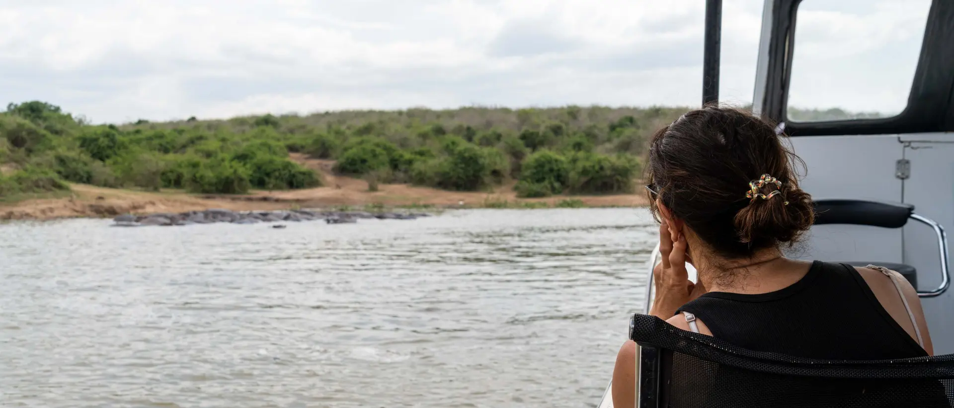 A person sitting on a boat, gazing thoughtfully at the water and surrounding greenery under a cloudy sky.