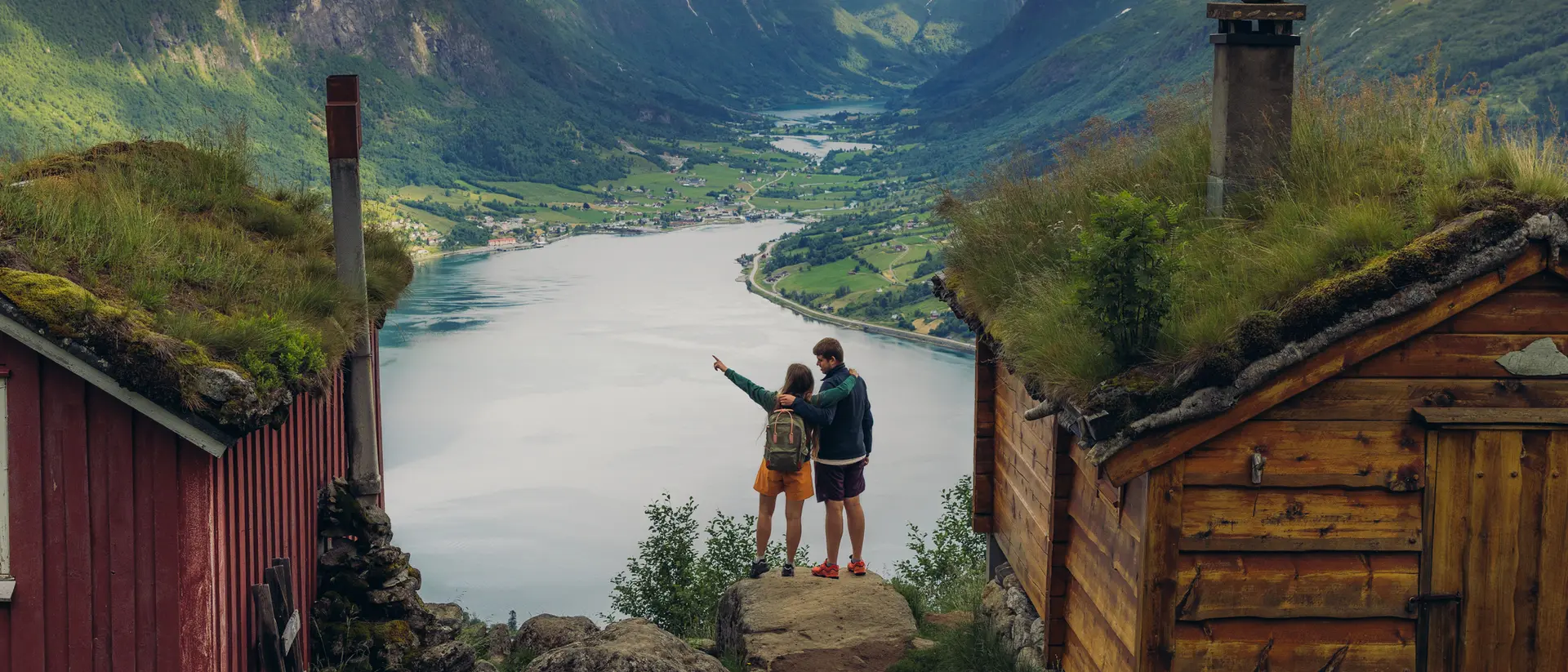 A couple stands between two wooden cabins, overlooking a scenic fjord surrounded by mountains.