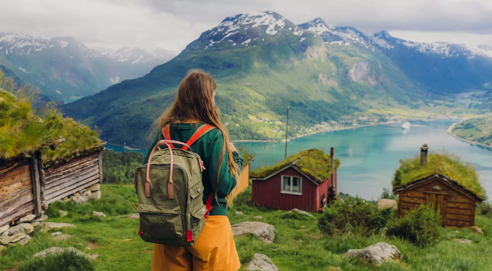 A person with a backpack stands on a hillside, overlooking a fjord and mountains with quaint wooden cabins.