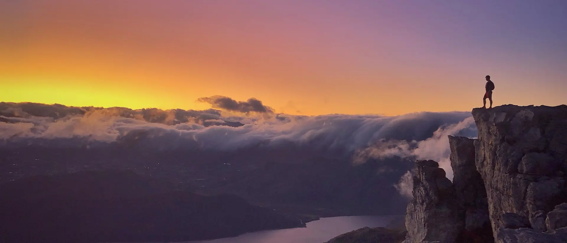 A person stands on a rocky ledge, overlooking a valley and lake at sunset, with colorful clouds in the sky.