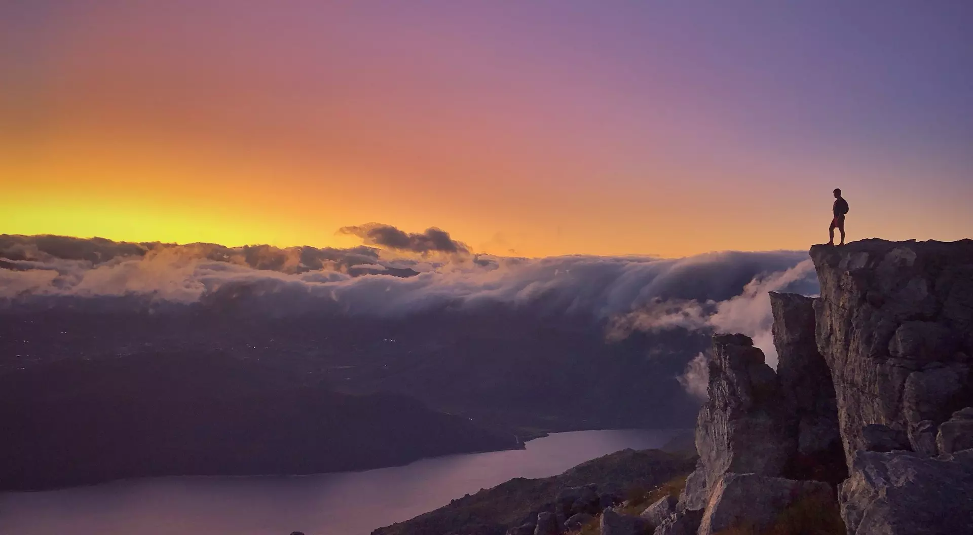 A person stands on a rocky ledge, overlooking a valley and lake at sunset, with colorful clouds in the sky.