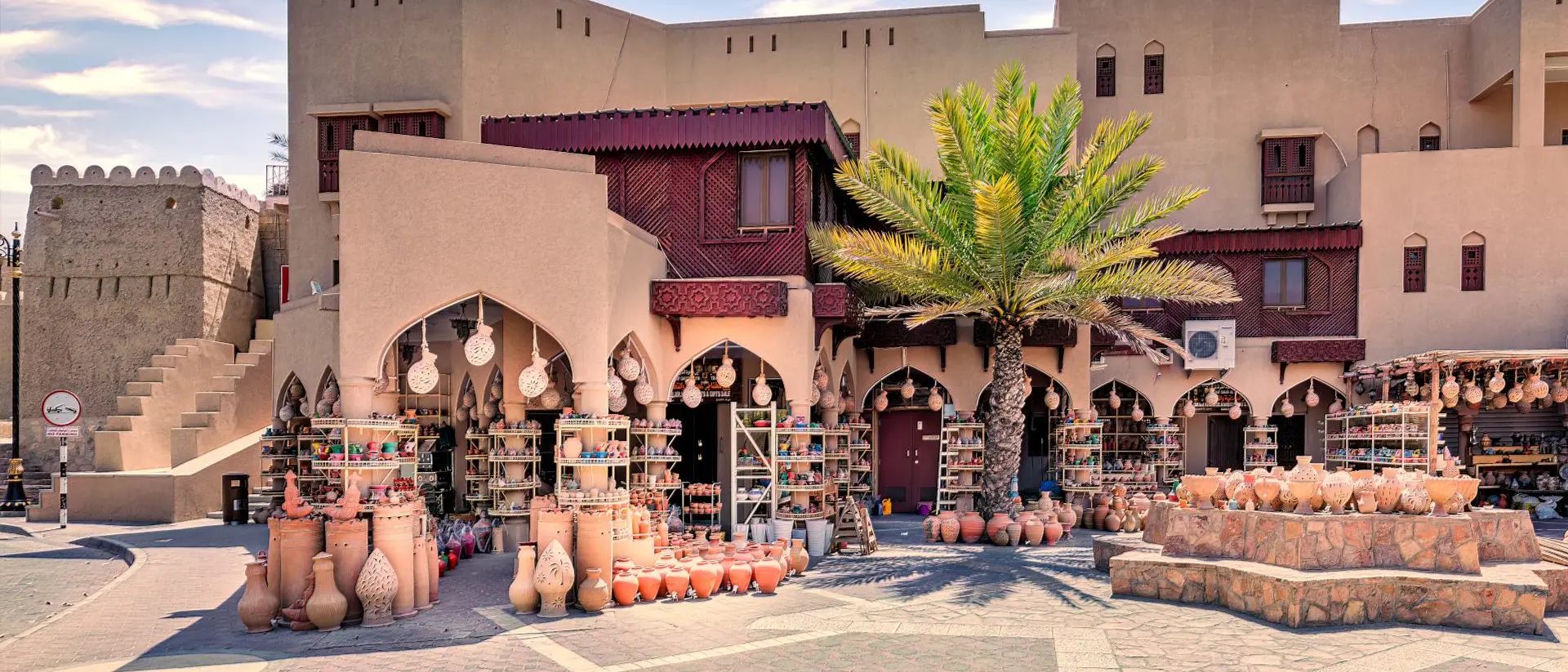 Market scene with clay pots for sale, palm trees, and traditional architecture under a blue sky.