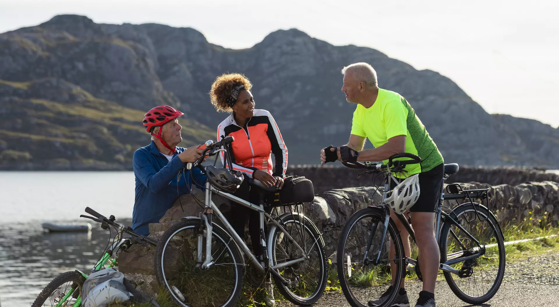 Three cyclists chat by a scenic lake, enjoying the outdoors in bright cycling gear against a mountainous backdrop.