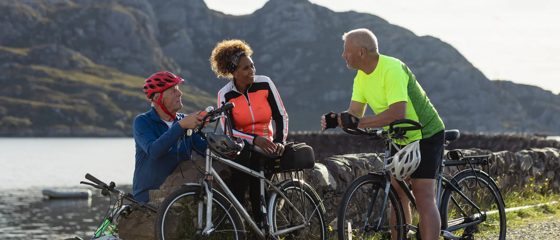 Three cyclists chat by a scenic lake, enjoying the outdoors in bright cycling gear against a mountainous backdrop.