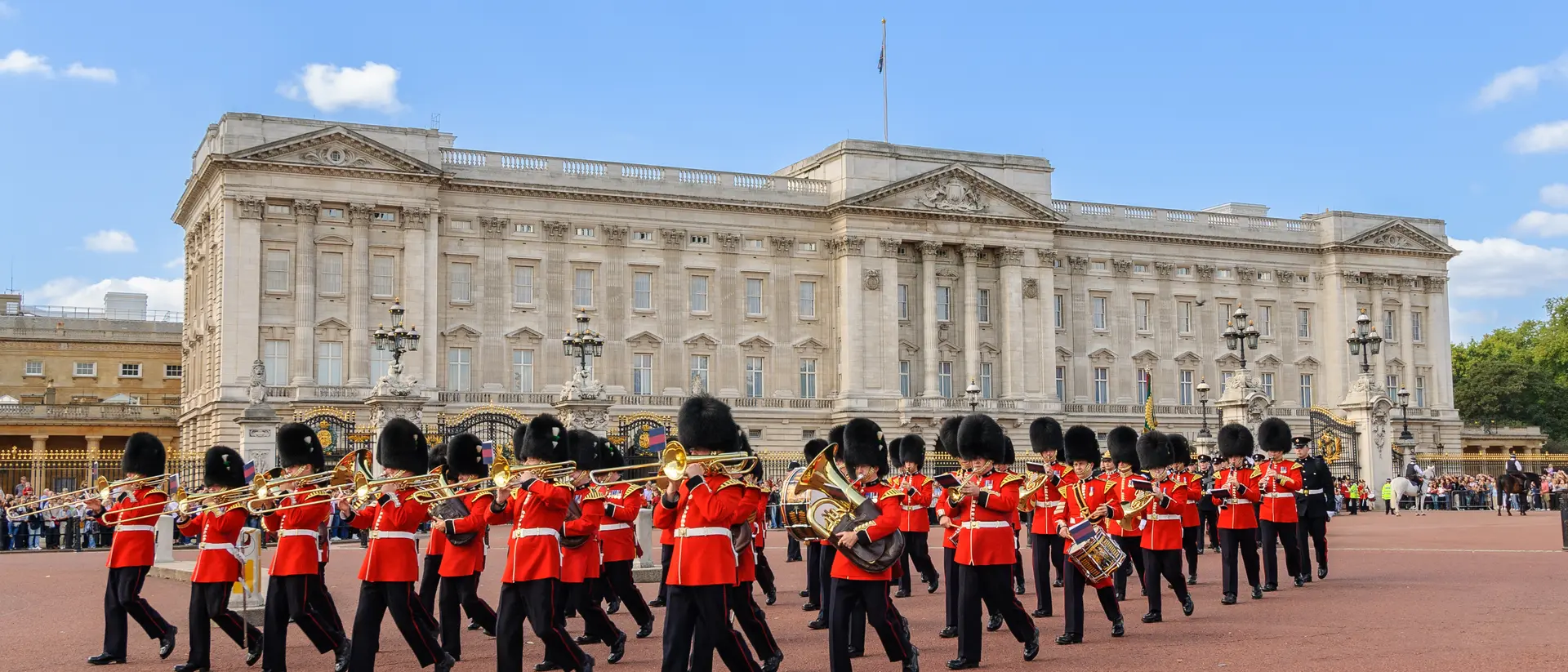 A marching band in red uniforms performs in front of Buckingham Palace under a blue sky.