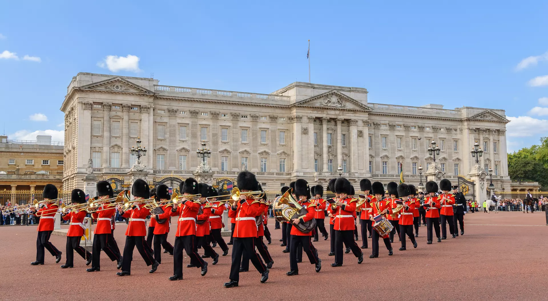 A marching band in red uniforms performs in front of Buckingham Palace under a blue sky.
