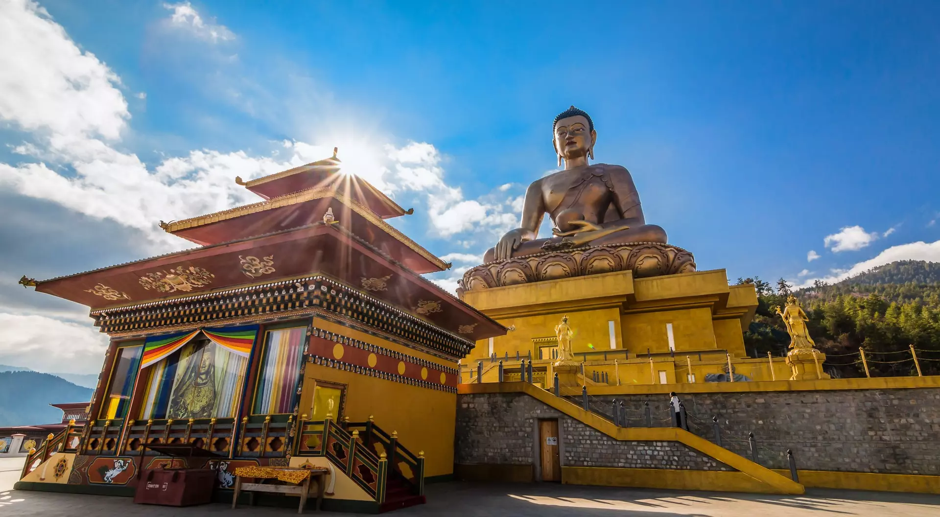 Golden Buddha statue beside a colorful temple under a bright sky, surrounded by mountains.