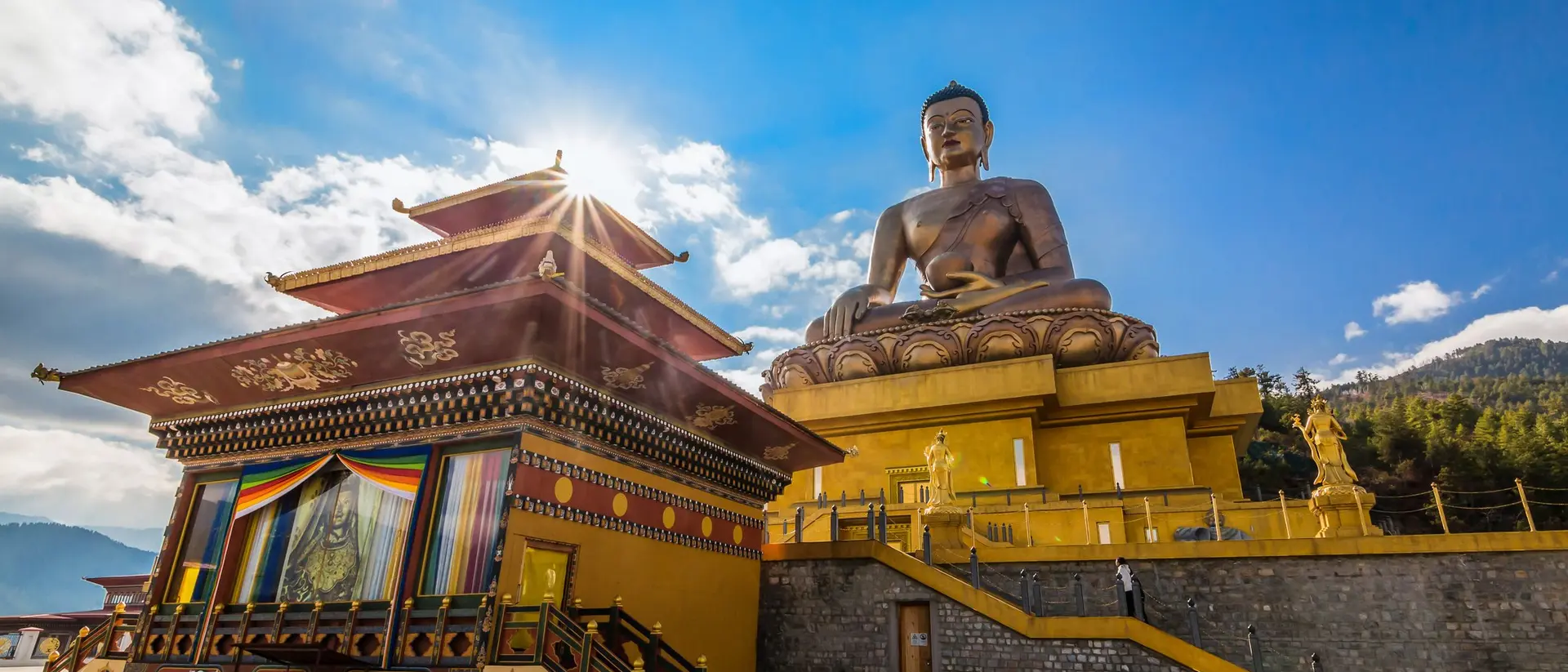 Golden Buddha statue beside a colorful temple under a bright sky, surrounded by mountains.