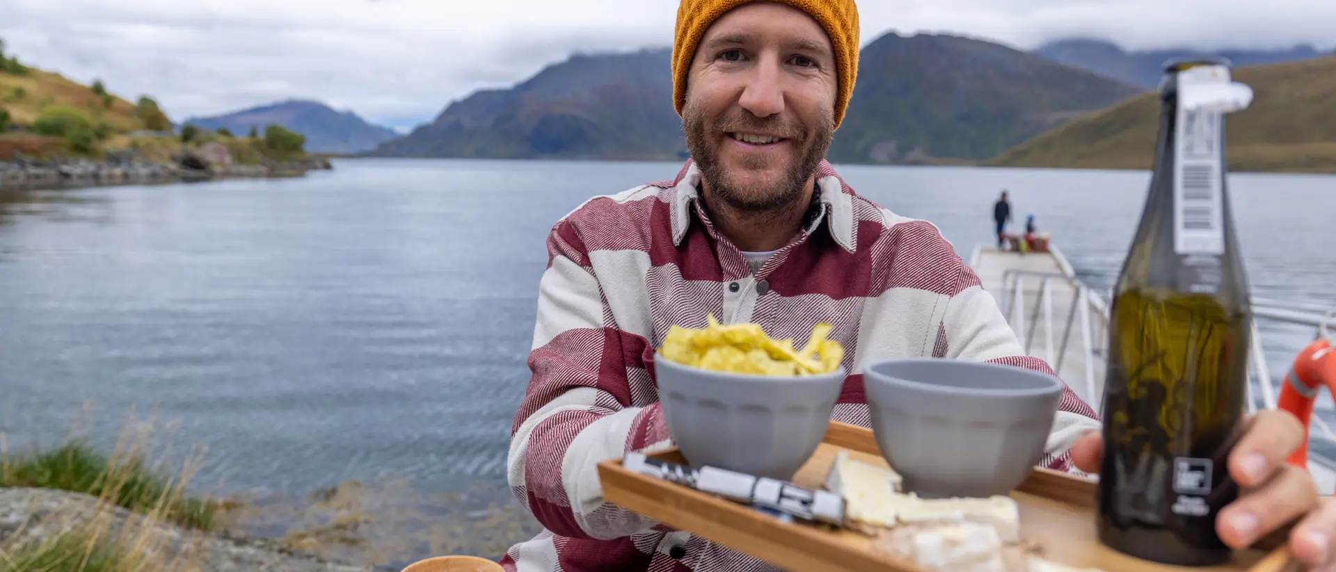 A smiling man in a striped shirt and orange beanie holds a serving tray with food and drinks by a calm lakeside.
