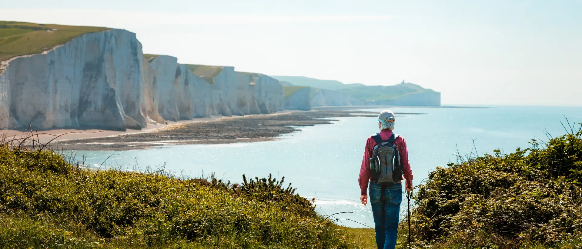Person walking along a grassy path with cliffs and ocean in the background on a sunny day.