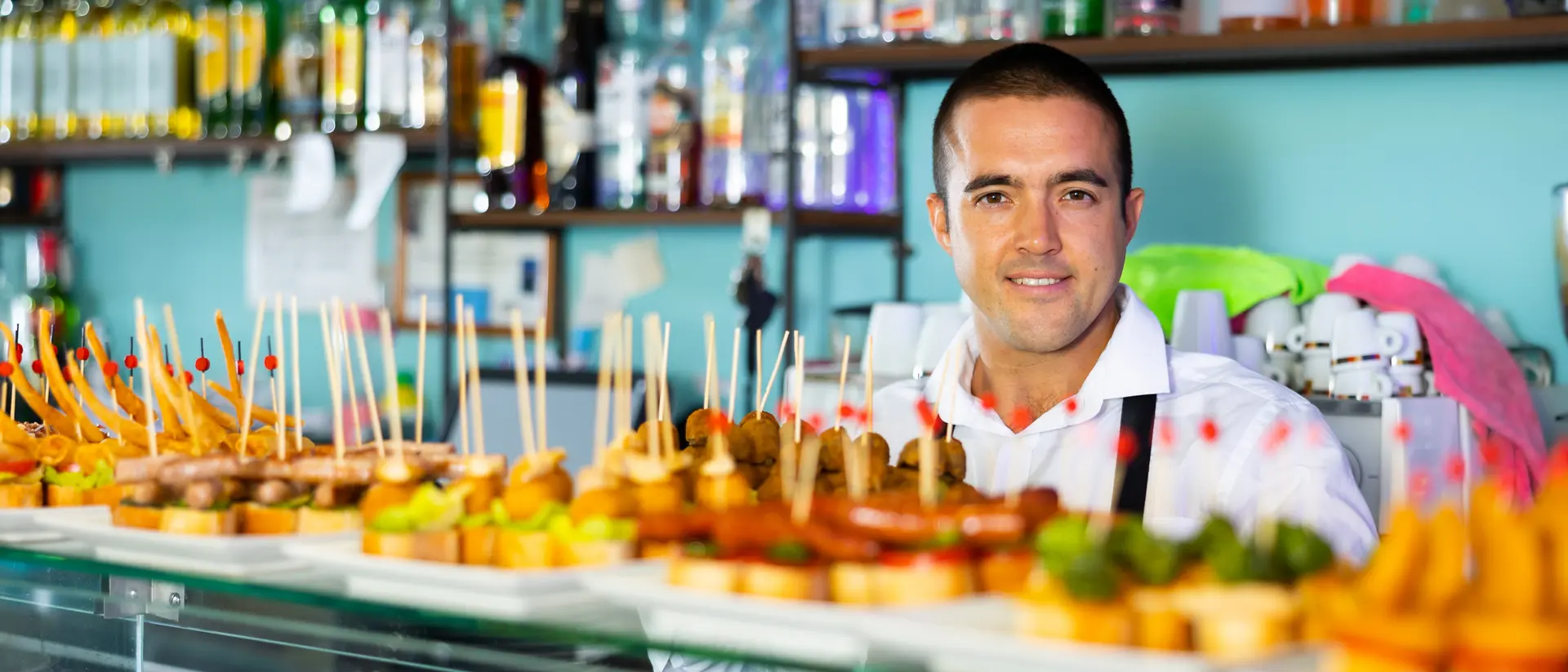 A smiling bartender stands behind a display of colorful tapas skewered with toothpicks, bottles of liquor visible in the background.