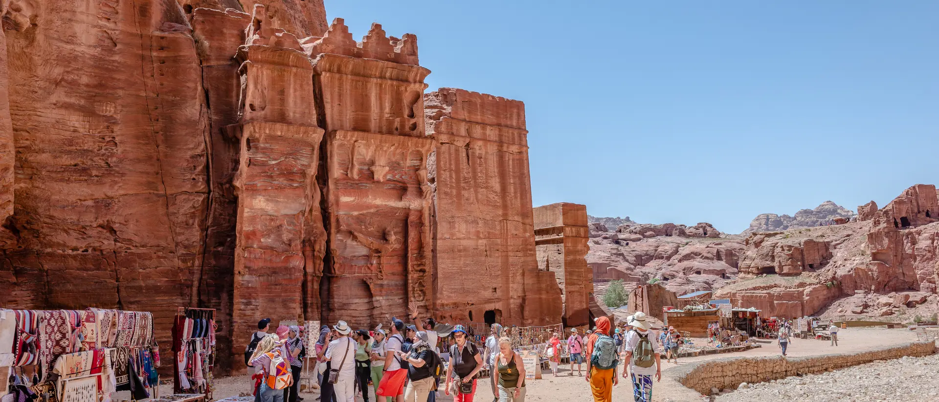 A bustling market near red rock formations, with tourists exploring and vendors selling colorful textiles and crafts.