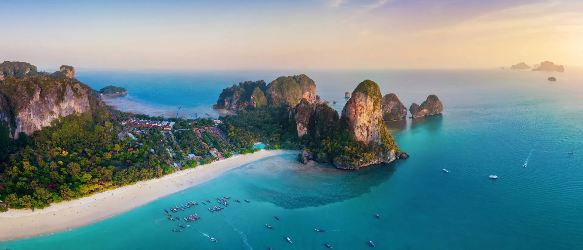 Aerial view of a tropical beach with clear blue waters, rocky cliffs, and colorful boats under a vibrant sky.
