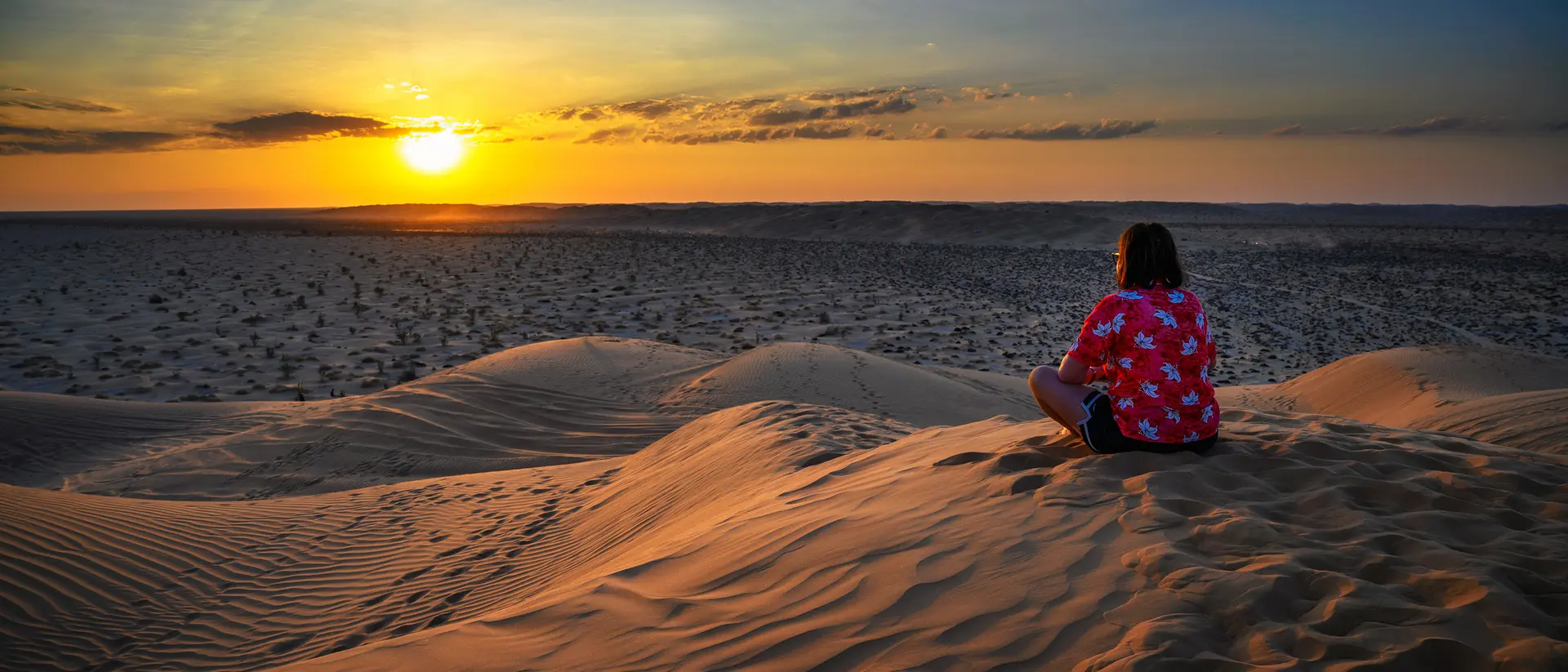 A person sitting on sand dunes, watching a colorful sunset against a vast desert landscape.