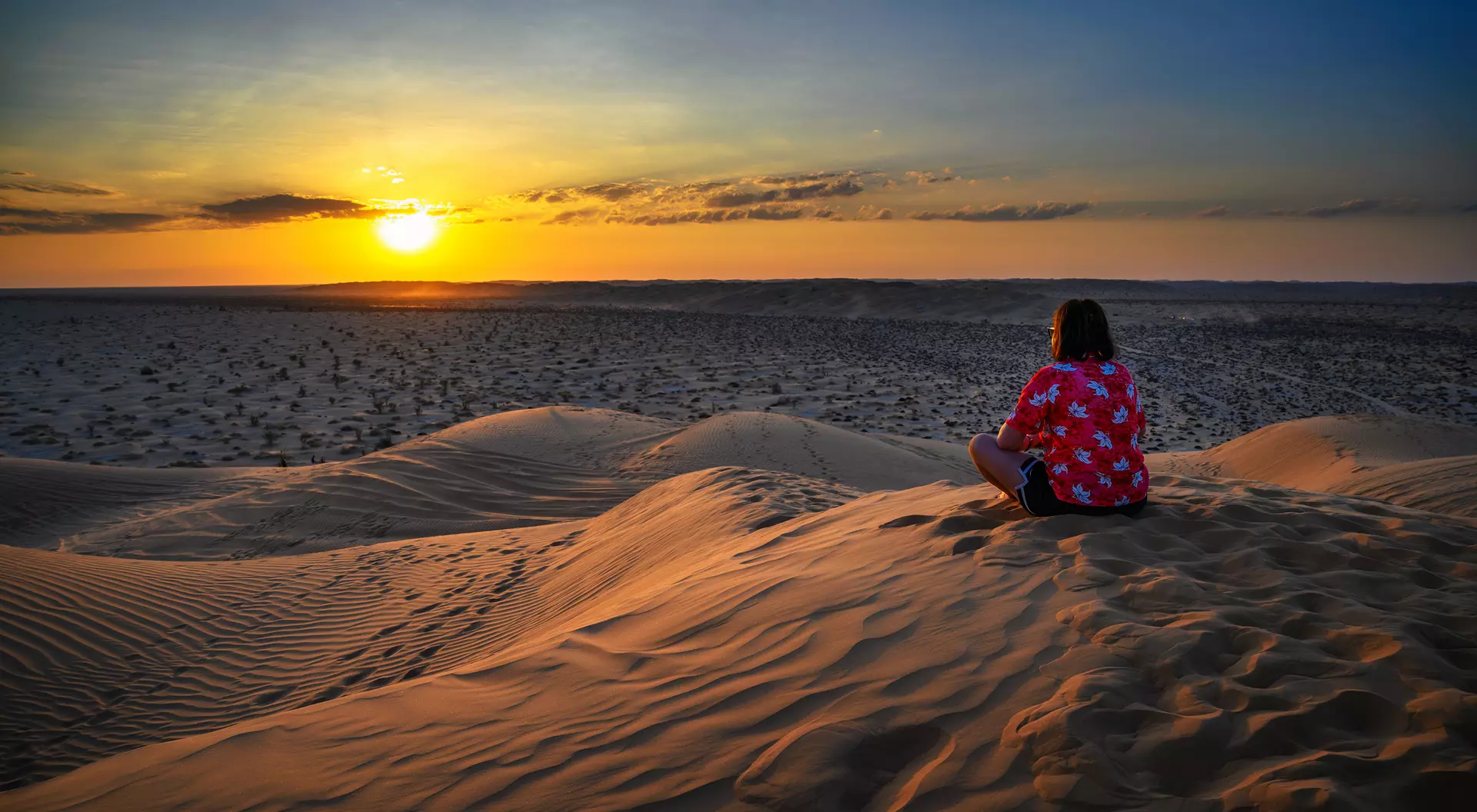 A person sitting on sand dunes, watching a colorful sunset against a vast desert landscape.