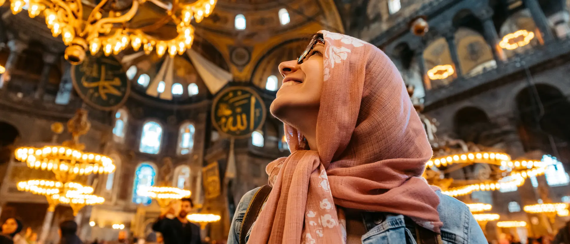 A woman in a hijab gazes upward in a historic, ornate building filled with chandeliers and people.