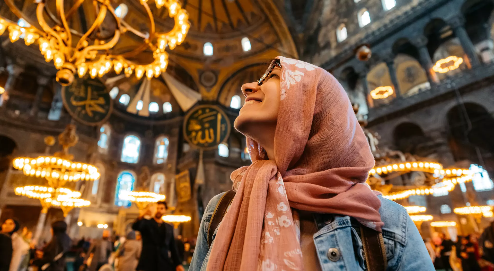 A woman in a hijab gazes upward in a historic, ornate building filled with chandeliers and people.