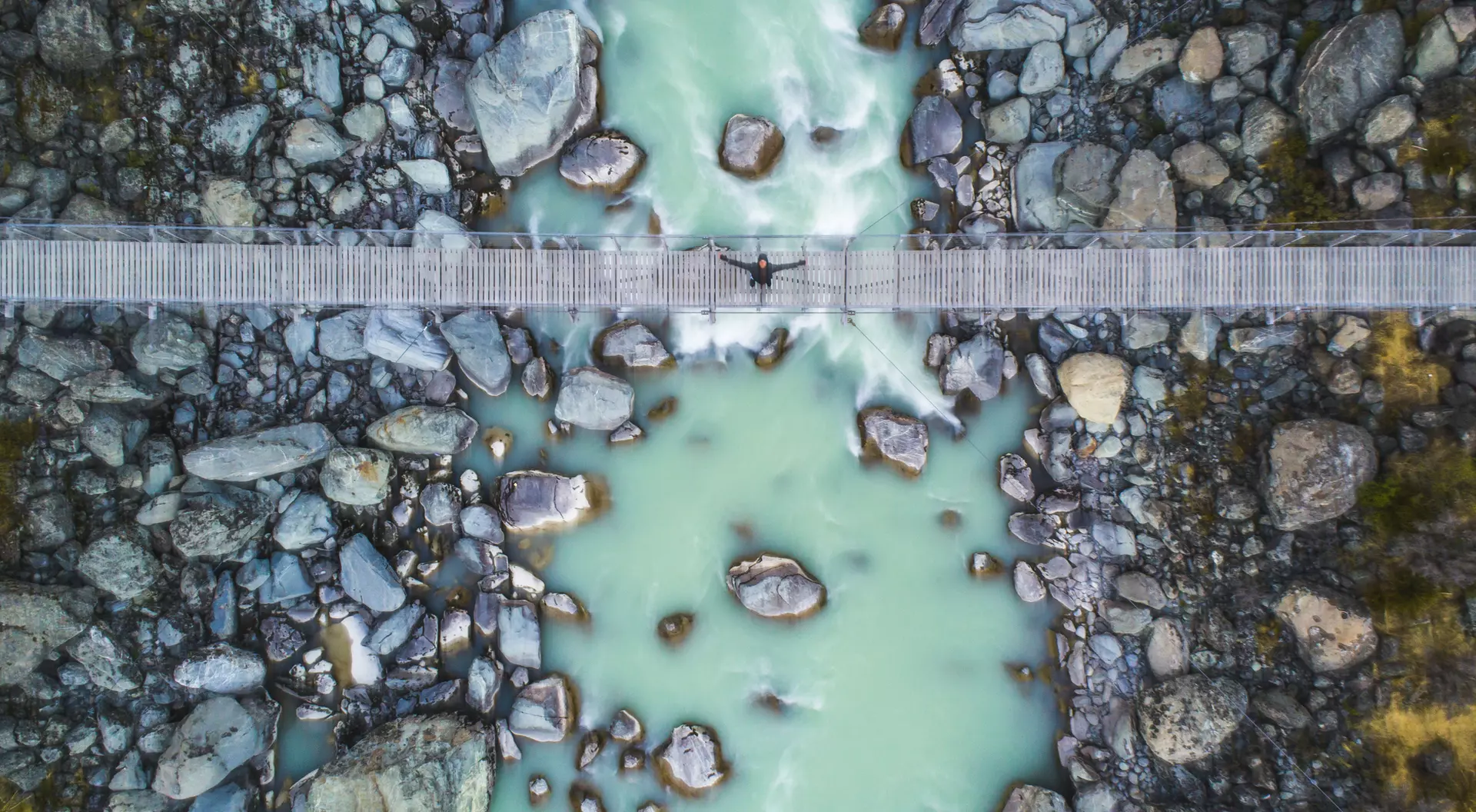 Aerial view of a river with turquoise water, surrounded by rocks, and a wooden bridge crossing over it.