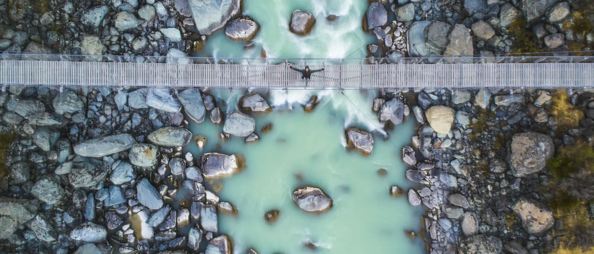 Aerial view of a river with turquoise water, surrounded by rocks, and a wooden bridge crossing over it.