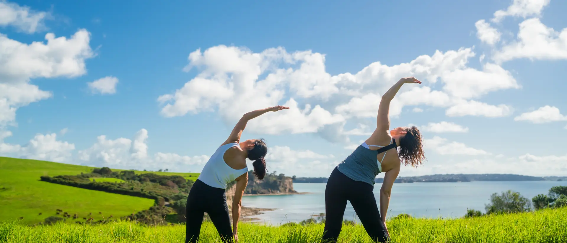 Two women stretching in a green field by a lake under a blue sky with fluffy clouds.