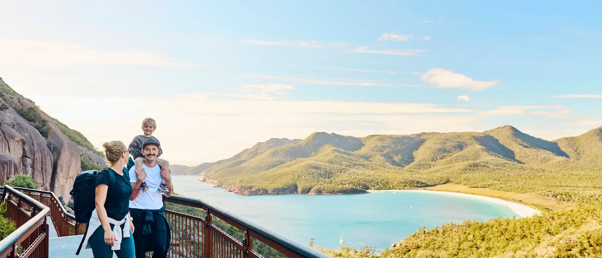 A family enjoys a scenic view from a coastal lookout, with hills and a beach in the background.
