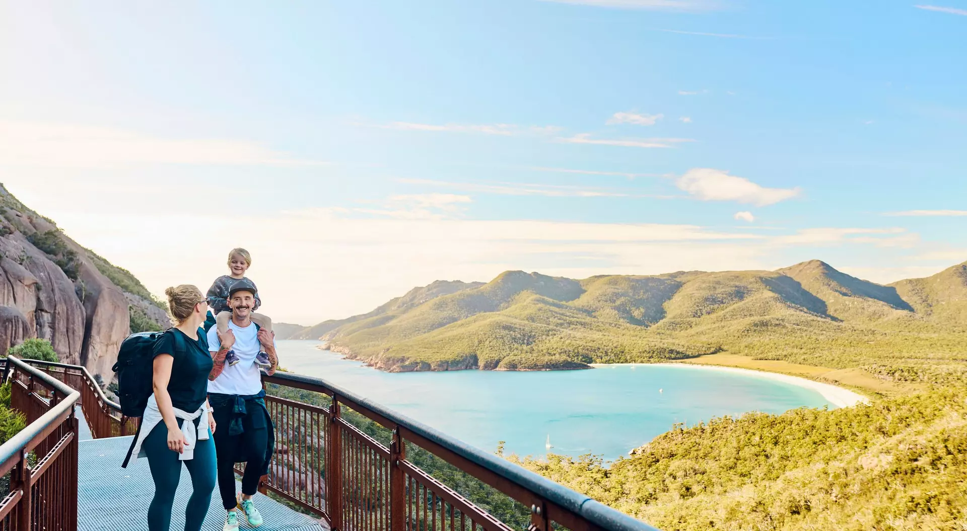 A family enjoys a scenic view from a coastal lookout, with hills and a beach in the background.