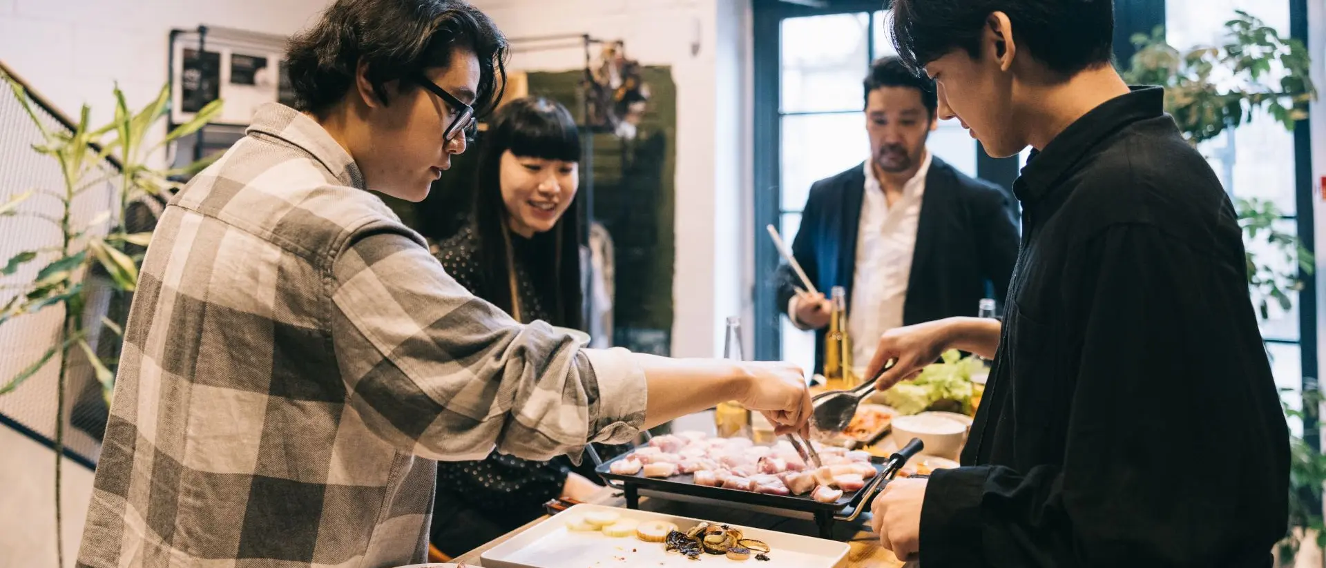 Group of friends enjoying a meal together, cooking at a table filled with various dishes and vibrant plants.