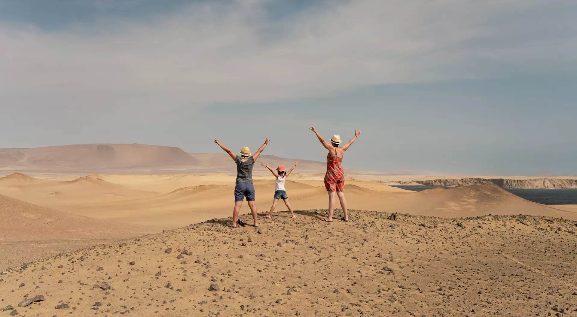 Three children stand on a desert hill, arms raised wide, with a vast sandy landscape and blue sky in the background.