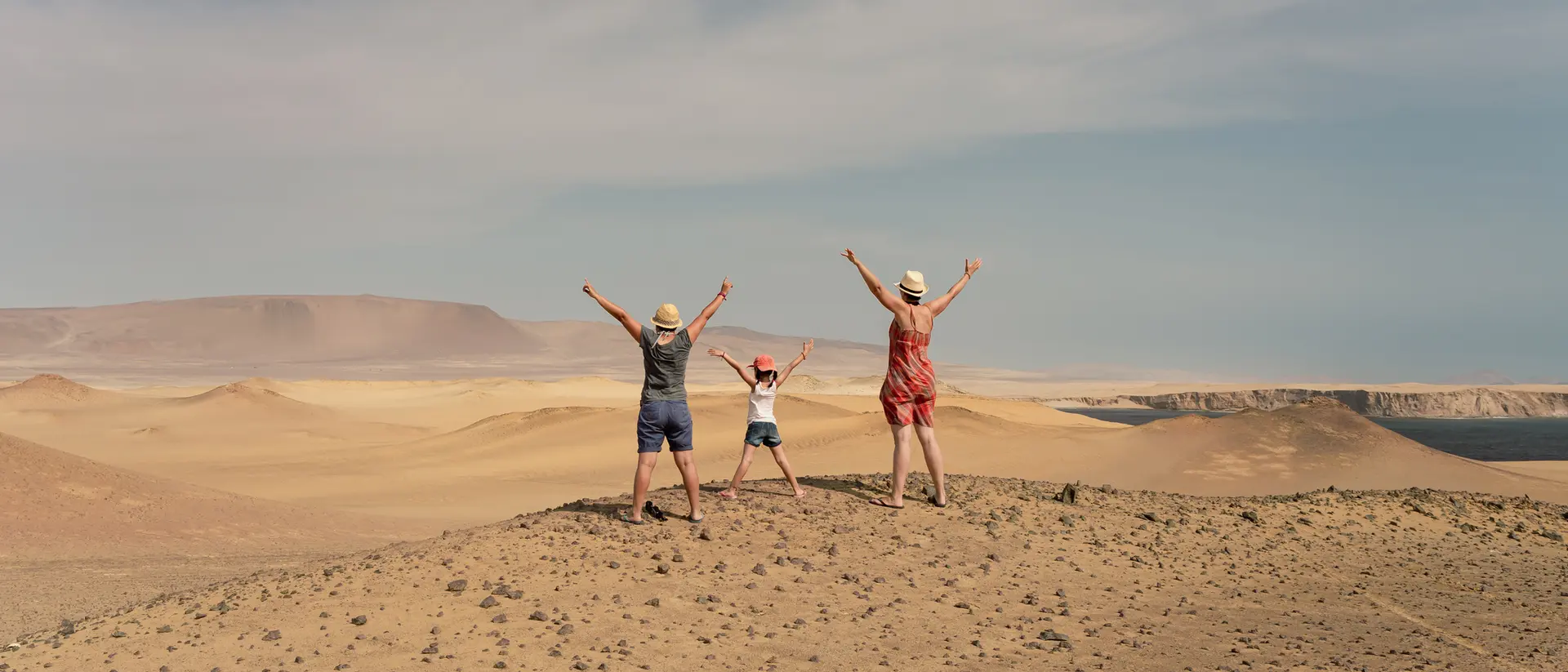 Three children stand on a desert hill, arms raised wide, with a vast sandy landscape and blue sky in the background.