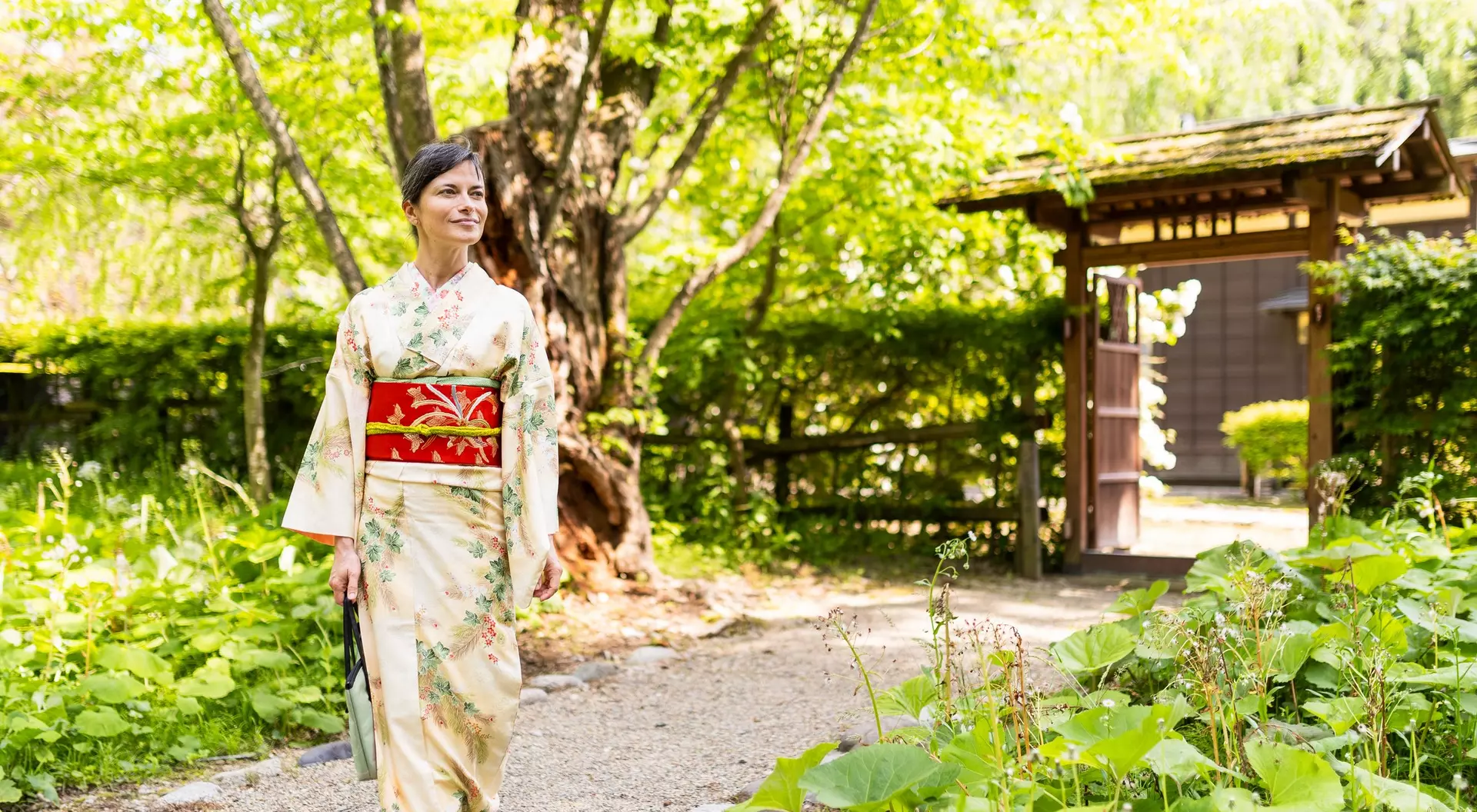 A woman in a traditional kimono walks through a lush, green garden near a wooden gate.