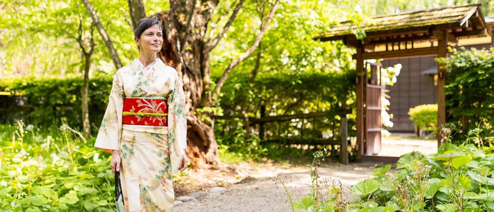 A woman in a traditional kimono walks through a lush, green garden near a wooden gate.