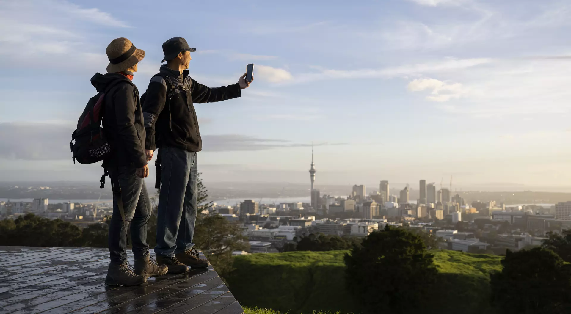 Two people take a selfie with a city skyline in the background during sunset, standing on a green hill.