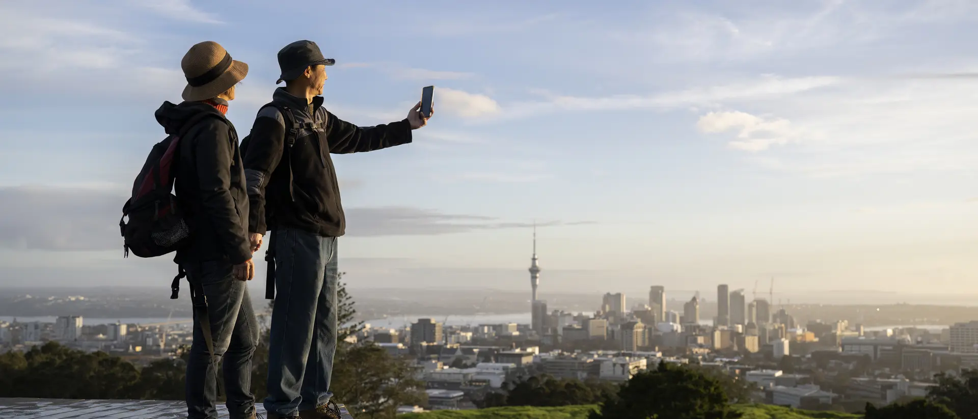 Two people take a selfie with a city skyline in the background during sunset, standing on a green hill.
