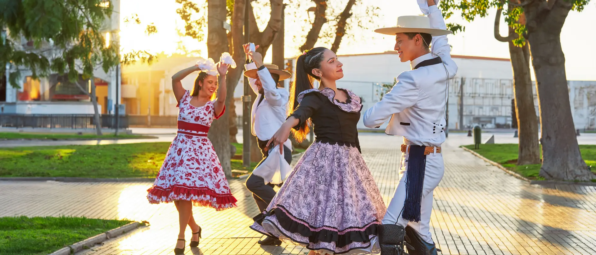 A couple dances in traditional attire outdoors, with more dancers in the background during sunset.