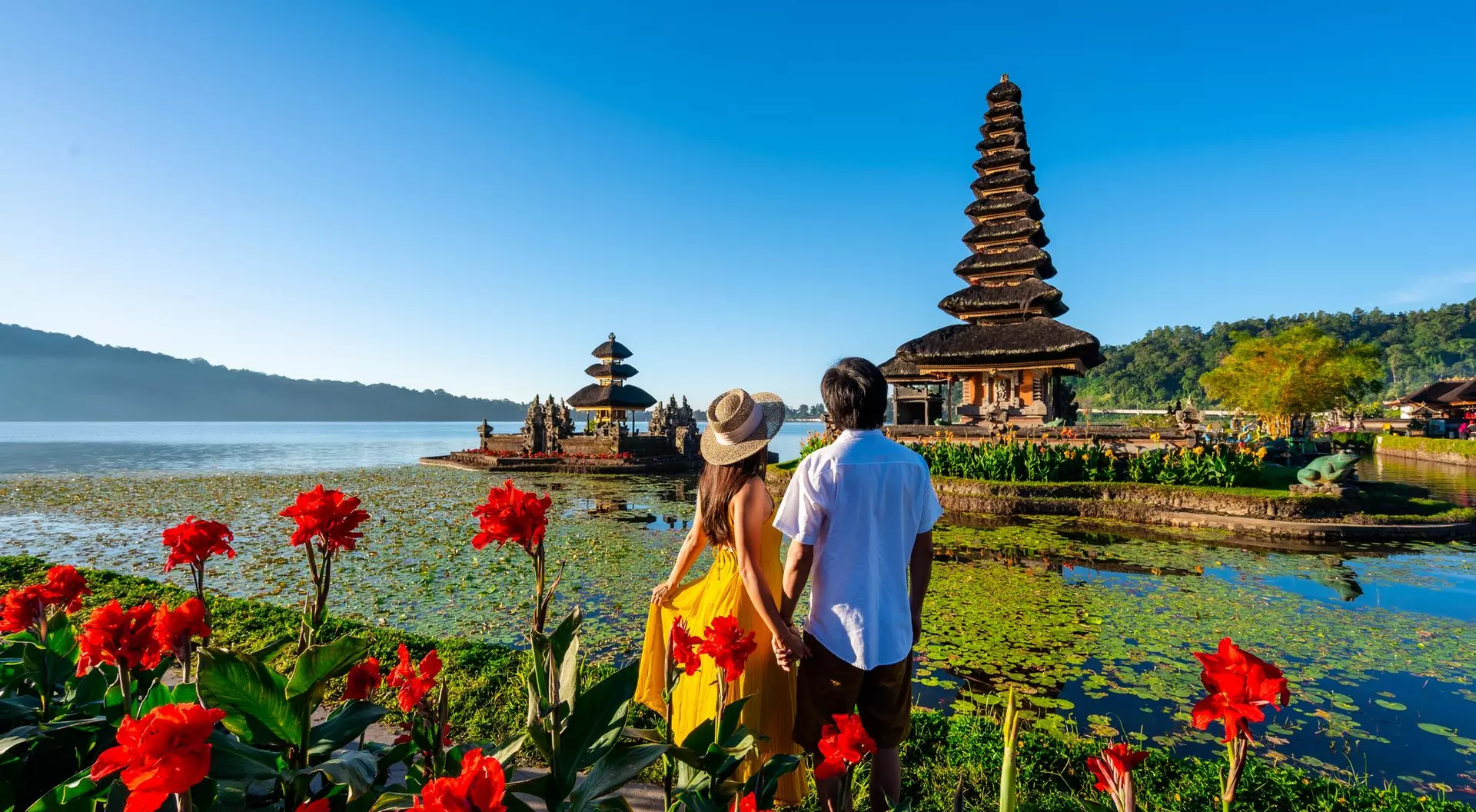 A couple holding hands by a lake, surrounded by vibrant flowers and a traditional pagoda in the background under a clear blue sky.