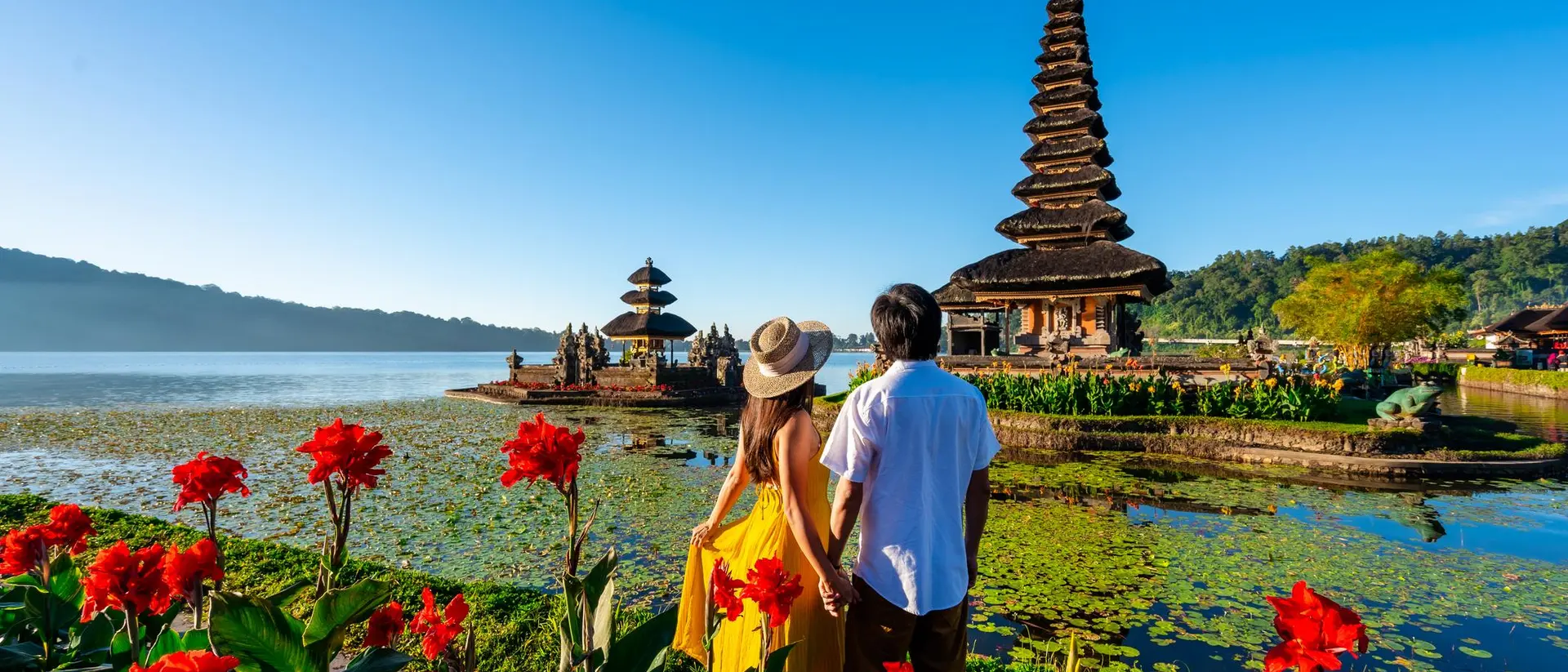 A couple holding hands by a lake, surrounded by vibrant flowers and a traditional pagoda in the background under a clear blue sky.