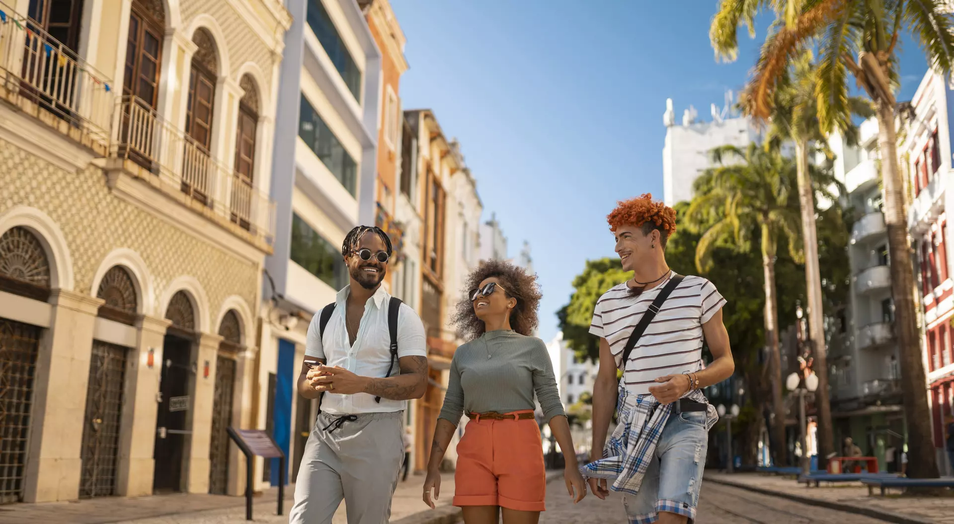 Three friends walk together on a sunny street lined with buildings and palm trees, enjoying their time outdoors.