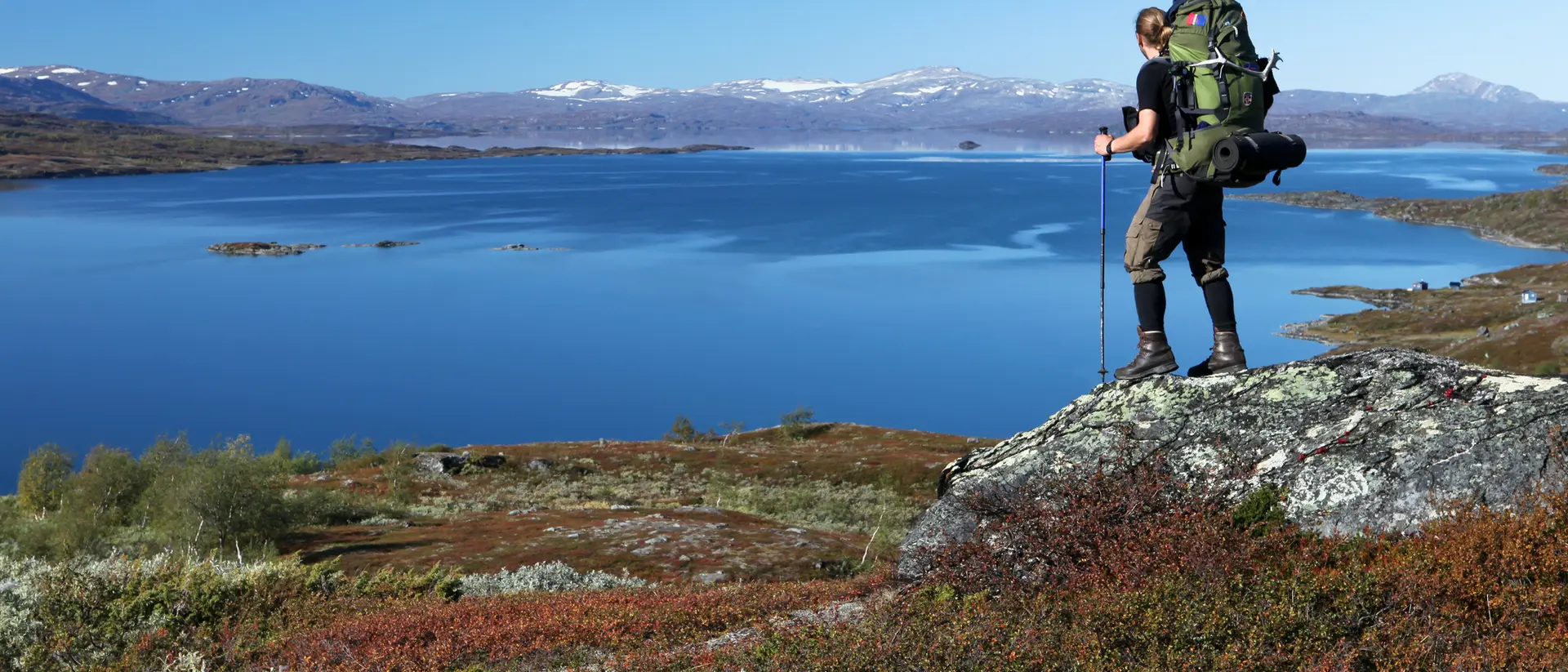 A hiker with a large backpack stands on a rock overlooking a calm lake and mountainous landscape under a clear blue sky.