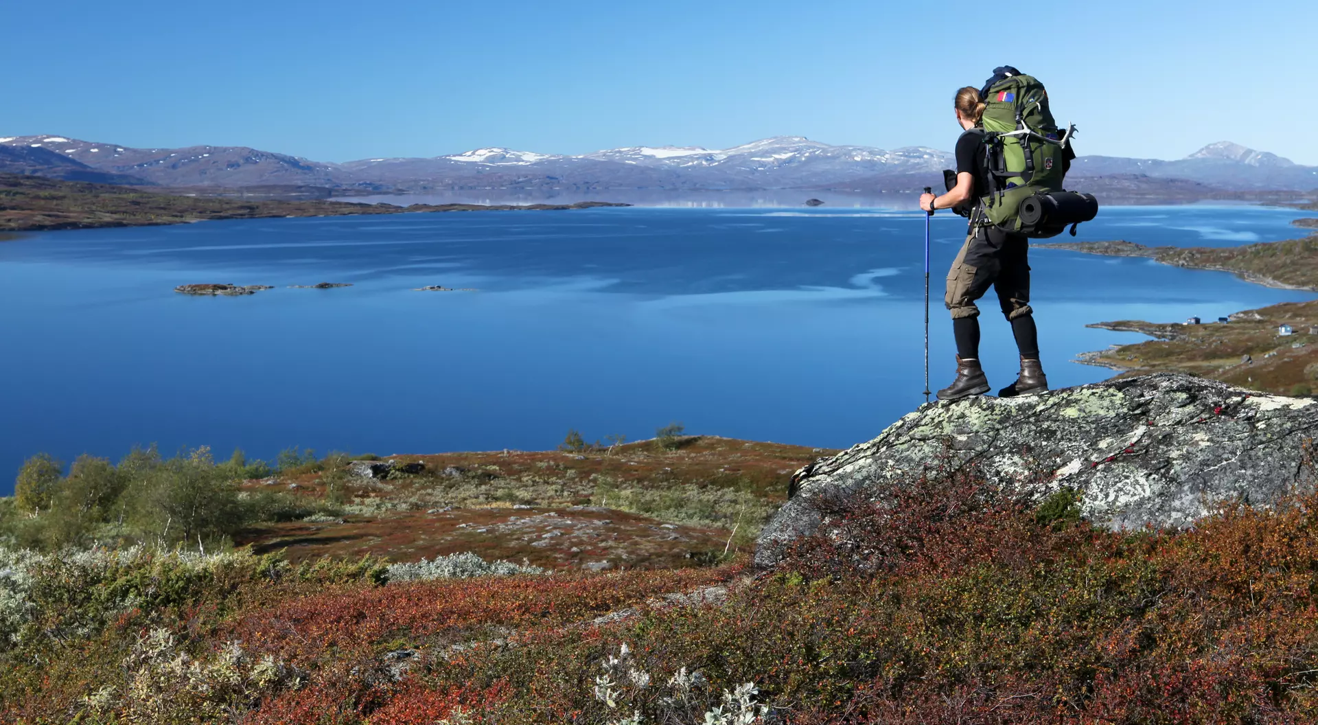 A hiker with a large backpack stands on a rock overlooking a calm lake and mountainous landscape under a clear blue sky.