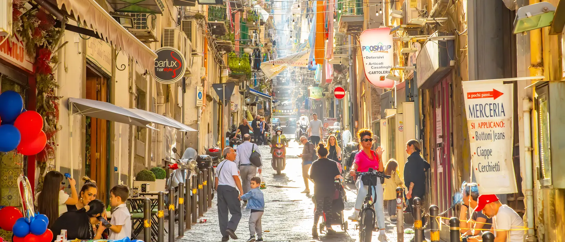 A bustling street scene with people walking, sitting at tables, and colorful decorations under bright sunshine.