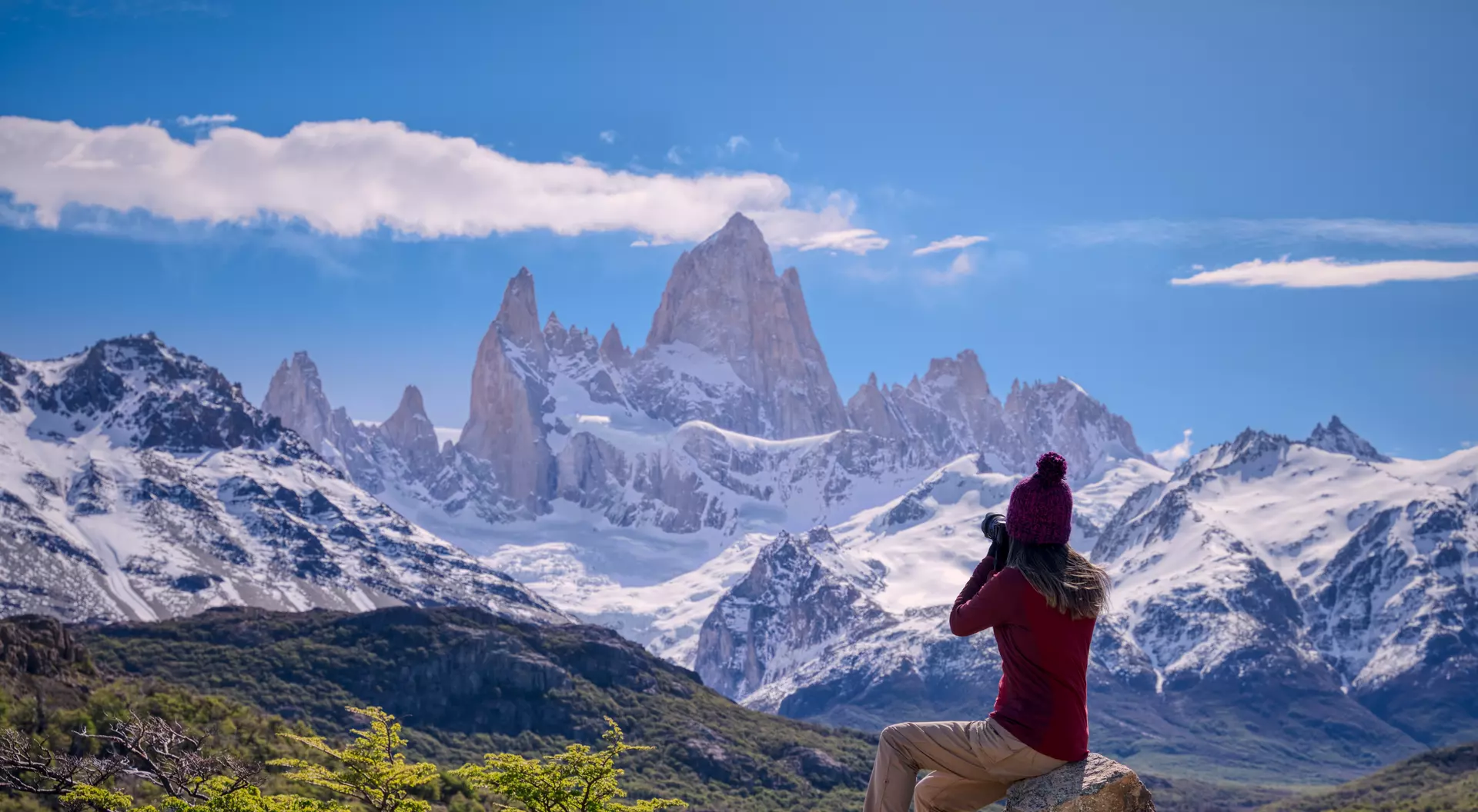 Person in a red sweater and hat photographs snow-capped mountains under a blue sky, seated on a rock.