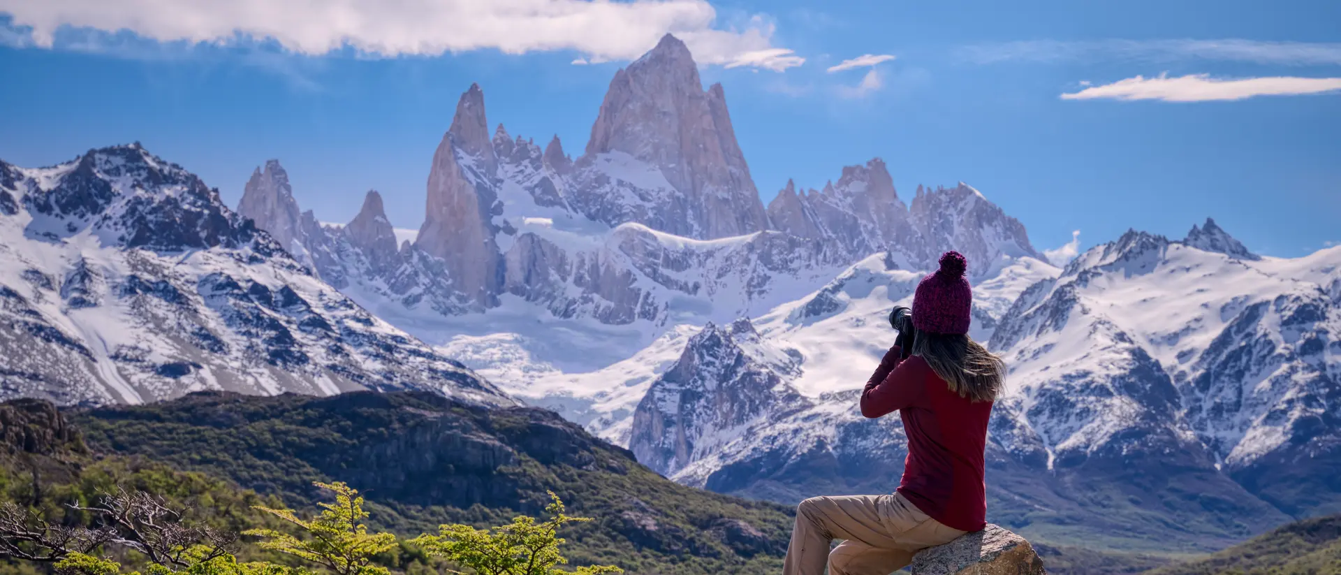 Person in a red sweater and hat photographs snow-capped mountains under a blue sky, seated on a rock.