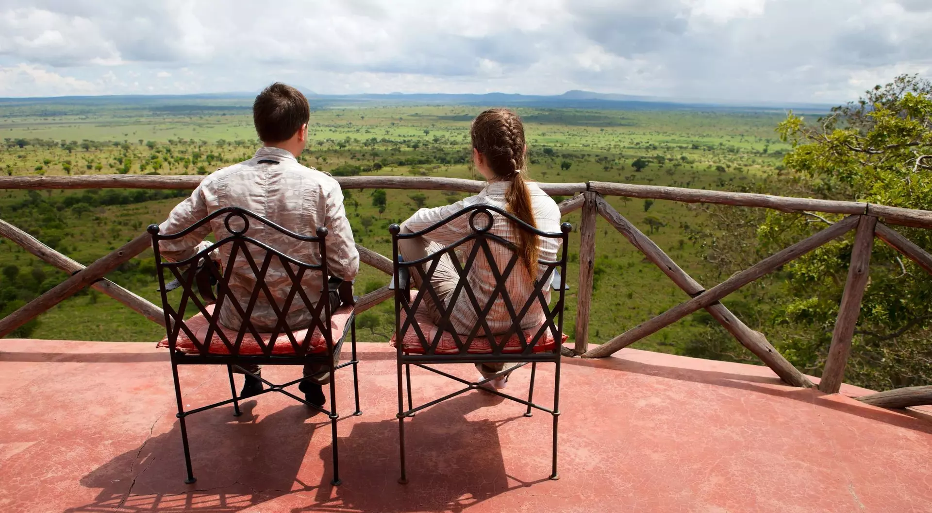 A couple sits in chairs on a balcony, gazing at a lush green landscape under a cloudy sky.