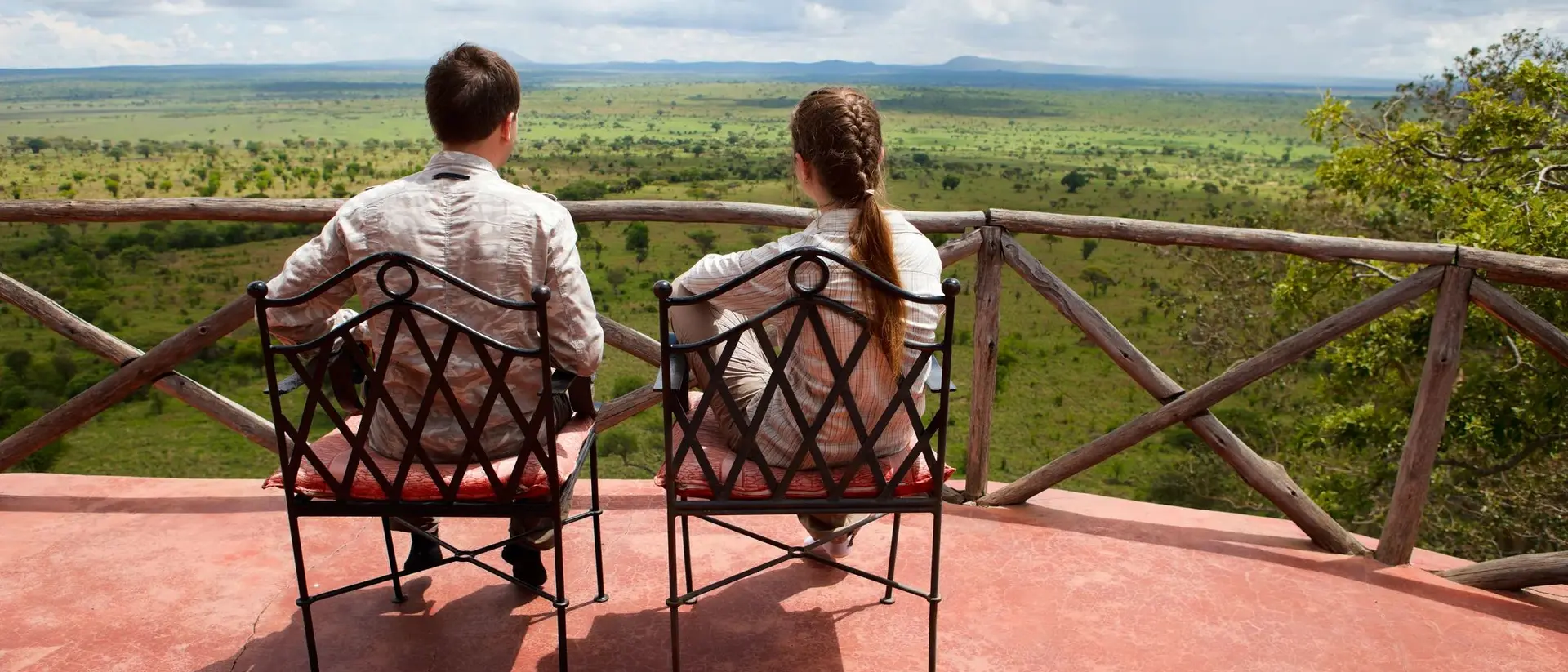 A couple sits in chairs on a balcony, gazing at a lush green landscape under a cloudy sky.