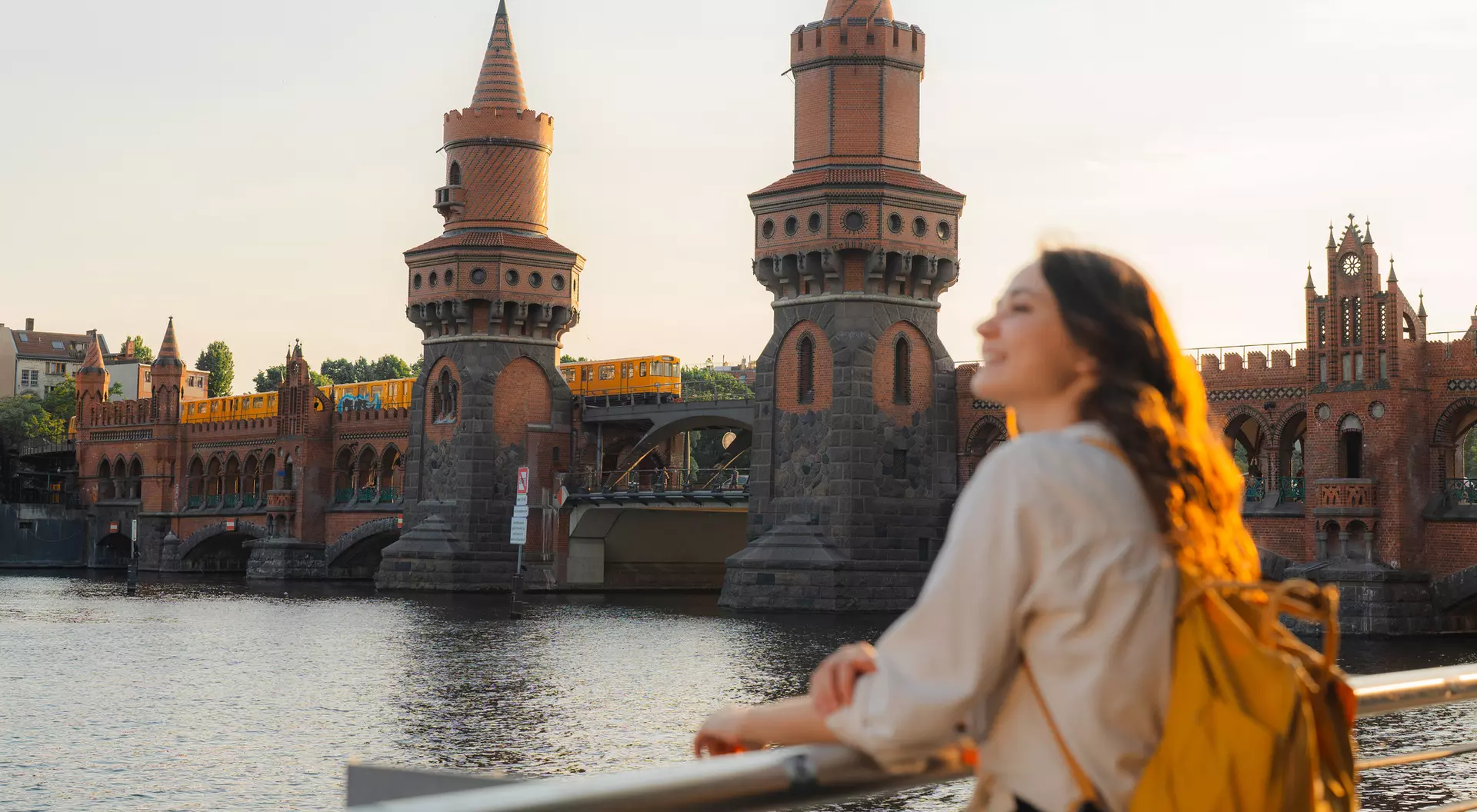Young woman with a backpack gazes at a historic bridge with towers above a river during sunset.