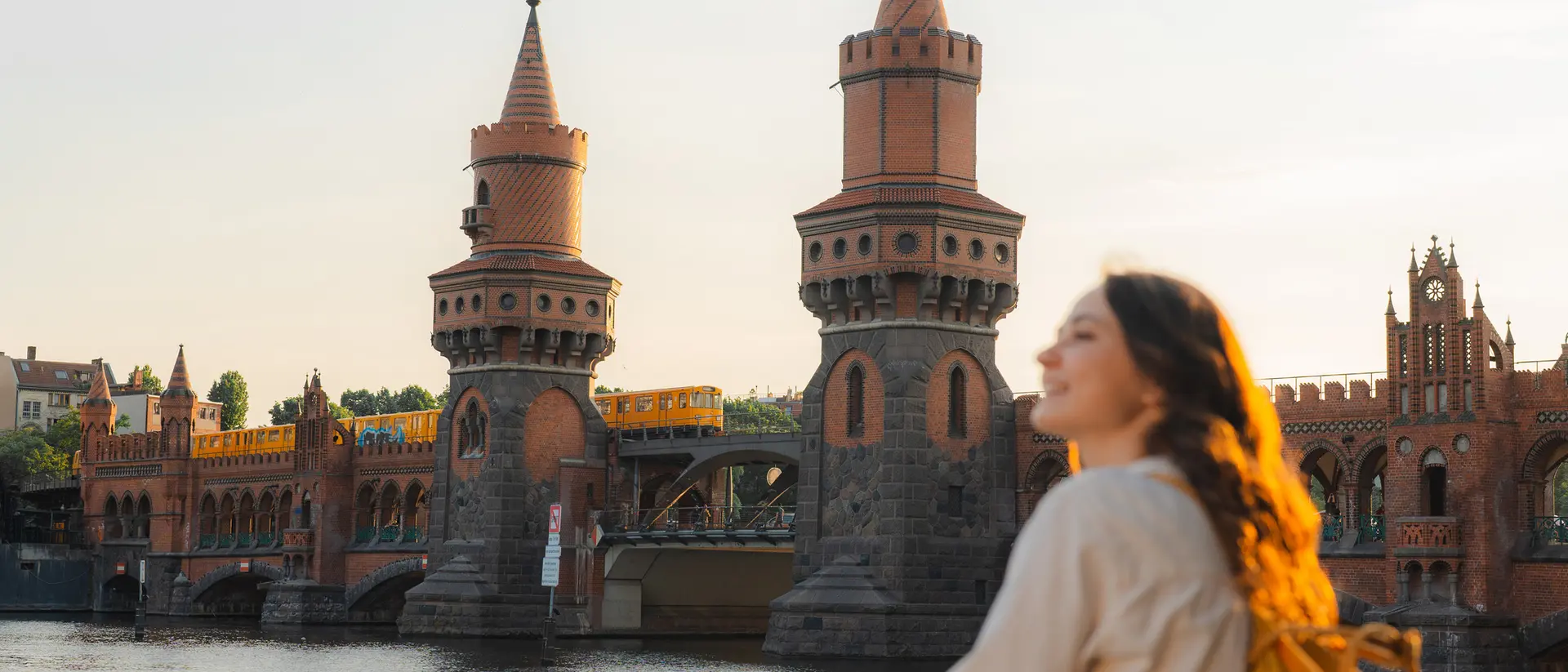 Young woman with a backpack gazes at a historic bridge with towers above a river during sunset.
