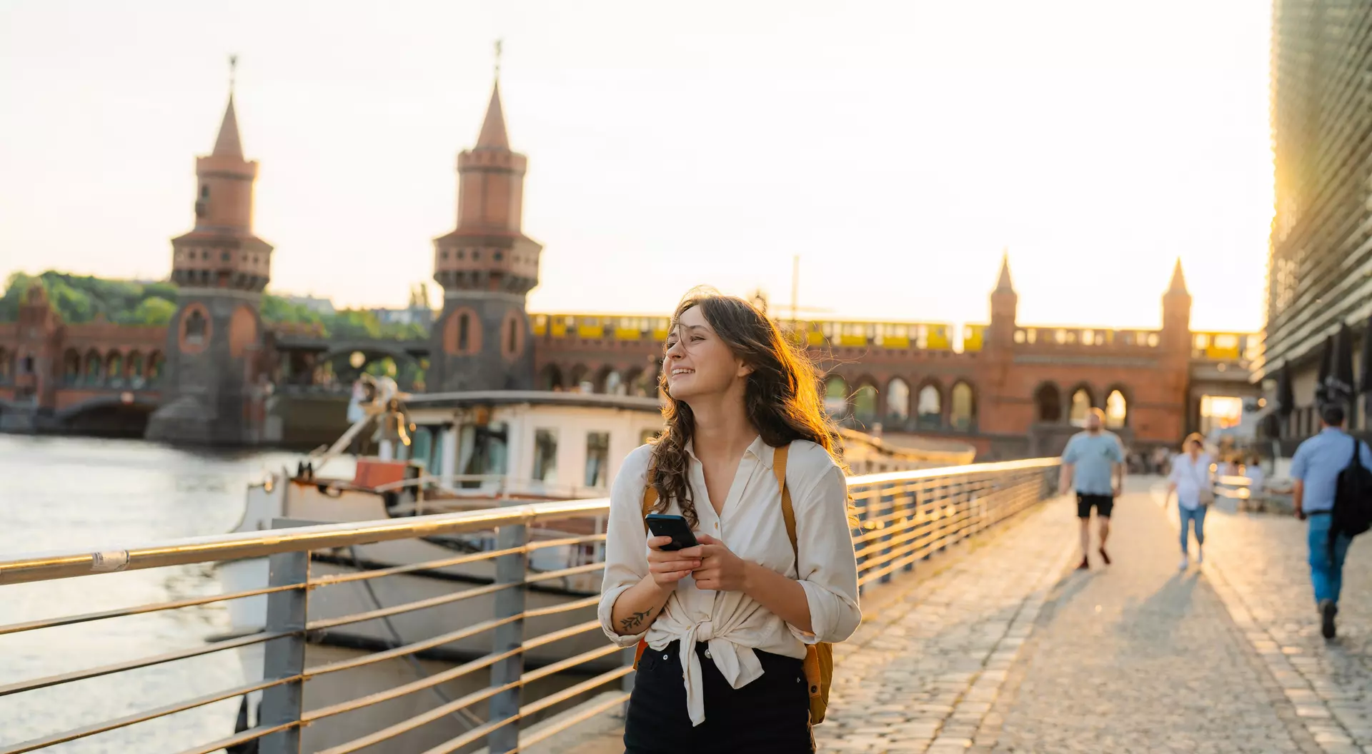 A woman stands on a riverside path, checking her phone, with a bridge and sunset in the background.
